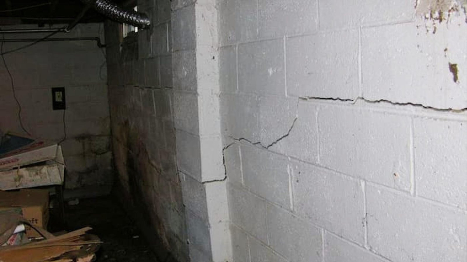 Dark basement corner with white-painted block wall showing long horizontal and diagonal cracks near electrical box, ductwork, and stored boxes.