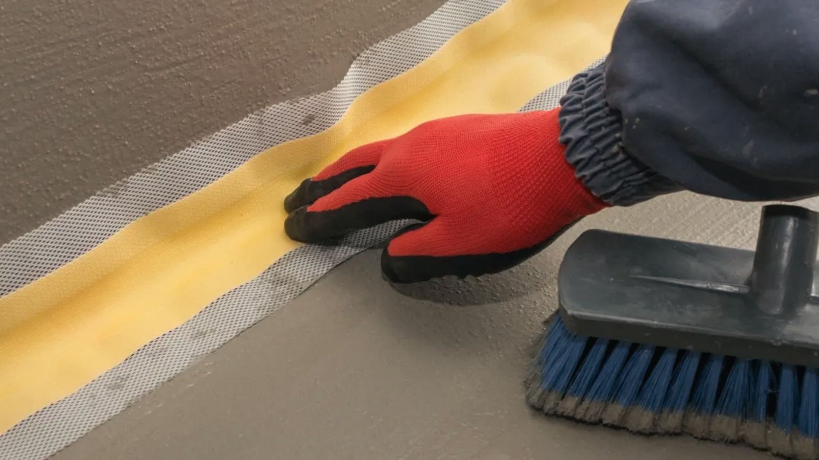 Gloved hand applying yellow waterproofing tape with mesh to basement floor joint for crack sealing