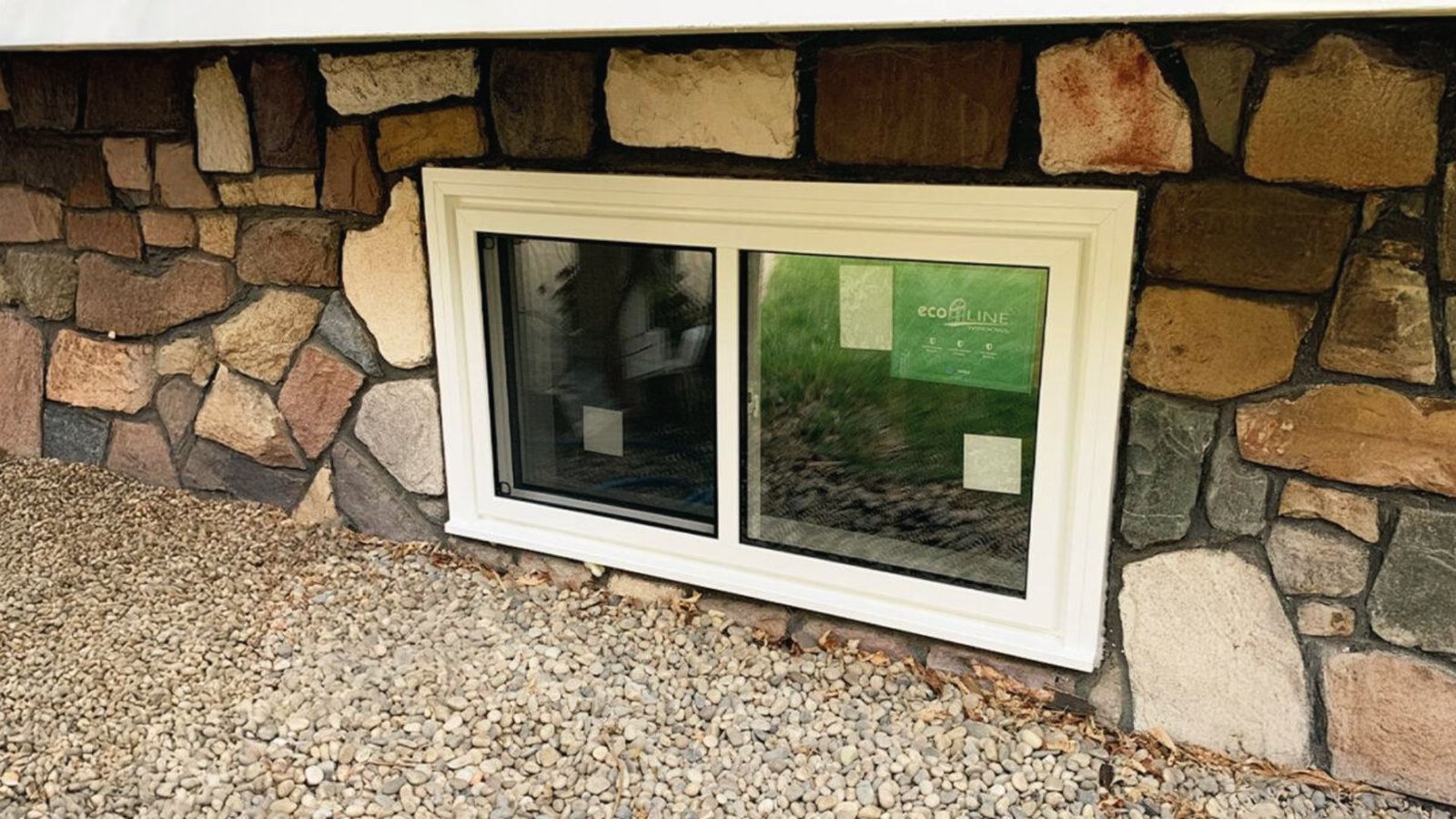White sliding basement window installed in stone wall above gravel bed for light and ventilation.