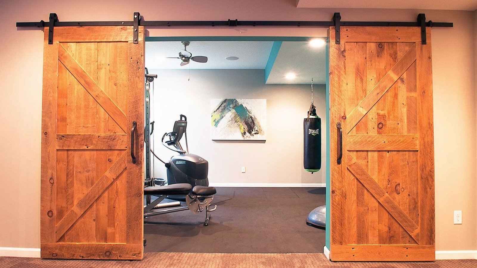 Basement home gym behind barn doors with punching bag, elliptical, weights and rubber flooring.