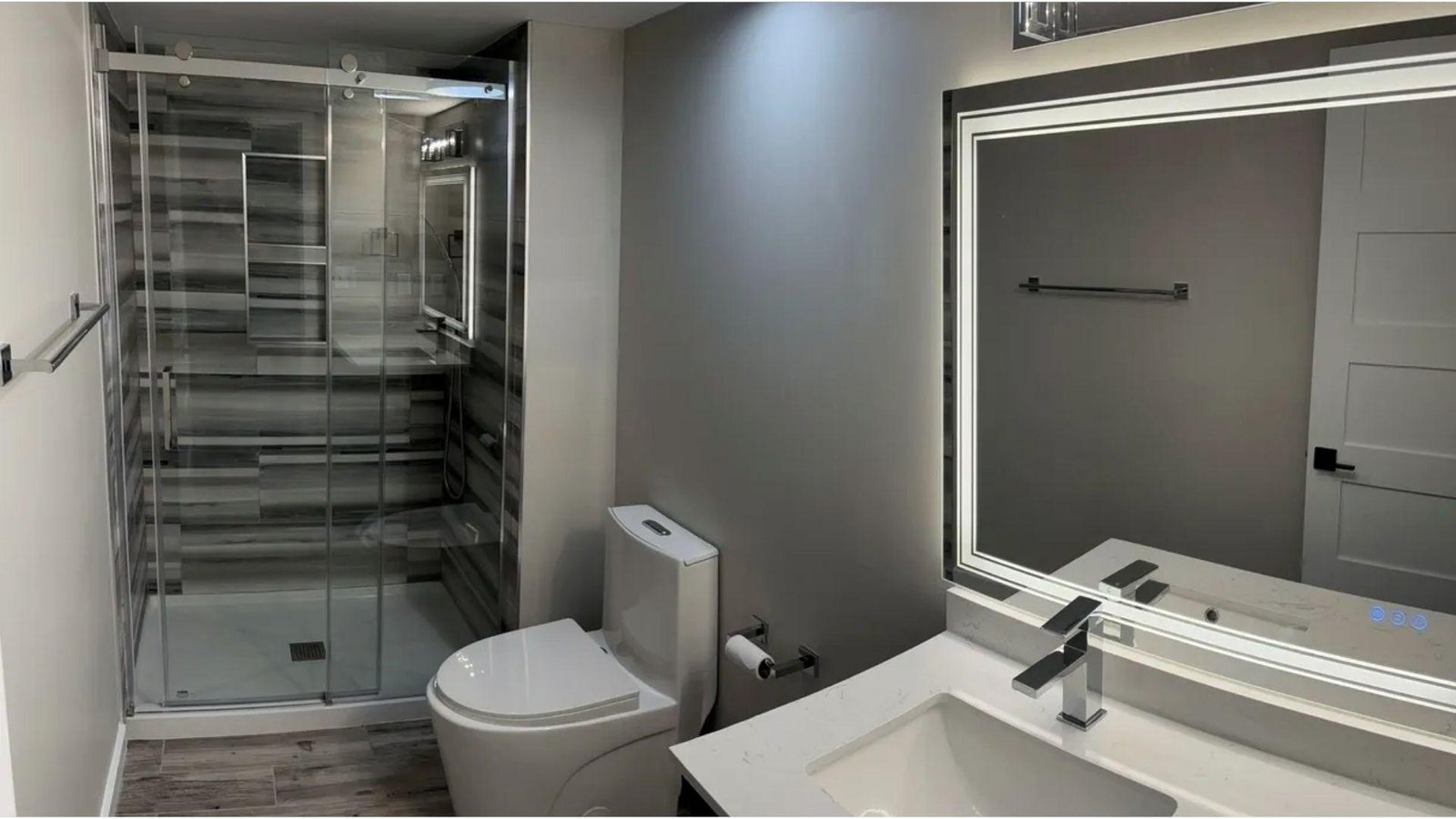 Modern basement bathroom with glass shower, toilet, and LED mirror