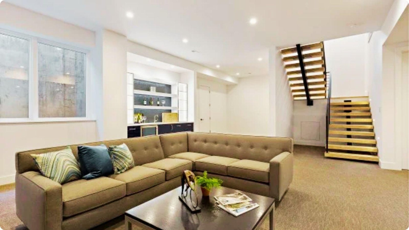English basement living room with a large tan sectional, built-in bar, and open-riser staircase leading to the main floor.