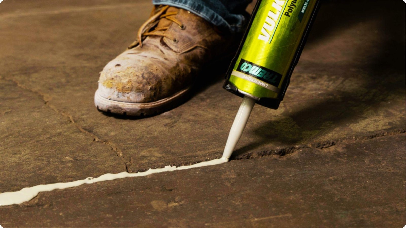 Close-up of a work boot and caulking gun applying white polyurethane sealant into a crack on a concrete floor.