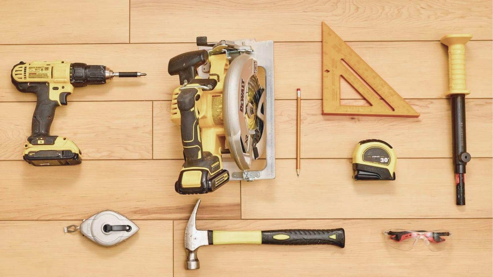 Flat lay of essential carpentry tools including a drill, circular saw, speed square, hammer, and tape measure on wood flooring.