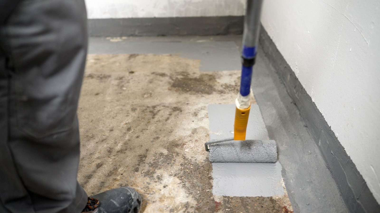 Worker using roller to apply gray waterproof sealant on basement concrete floor to prevent water damage