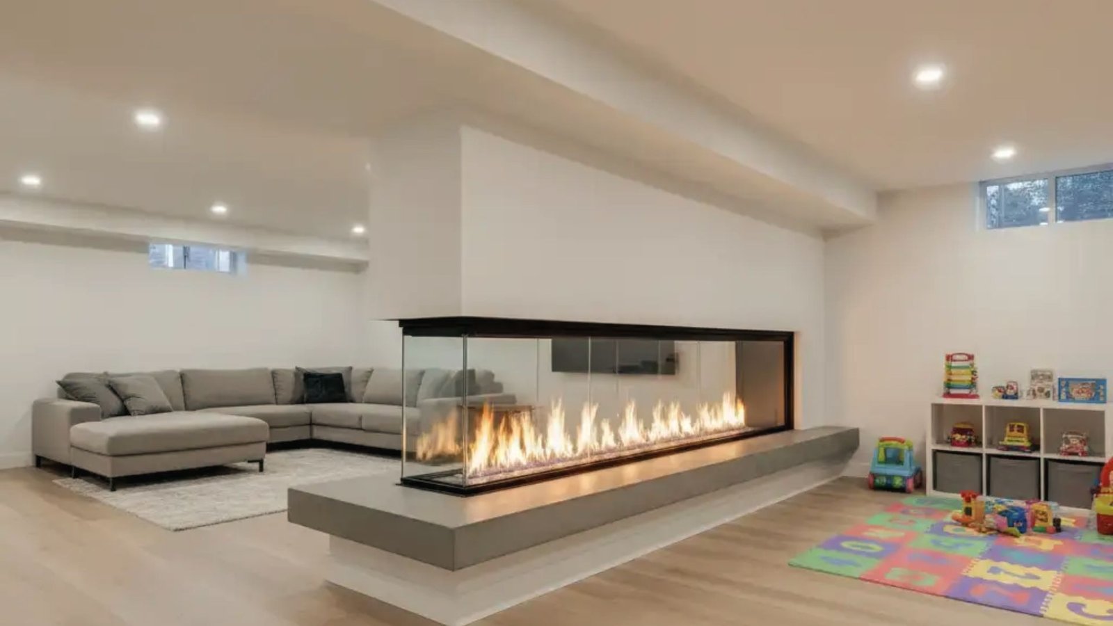 Three-sided linear gas fireplace in basement playroom with gray sectional and toy storage.