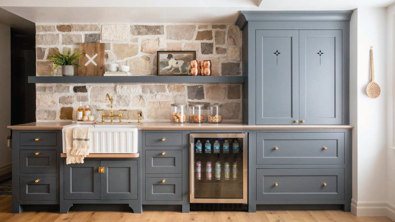 Modern farmhouse basement wet bar with stone wall, farmhouse sink, blue cabinets and mini fridge.