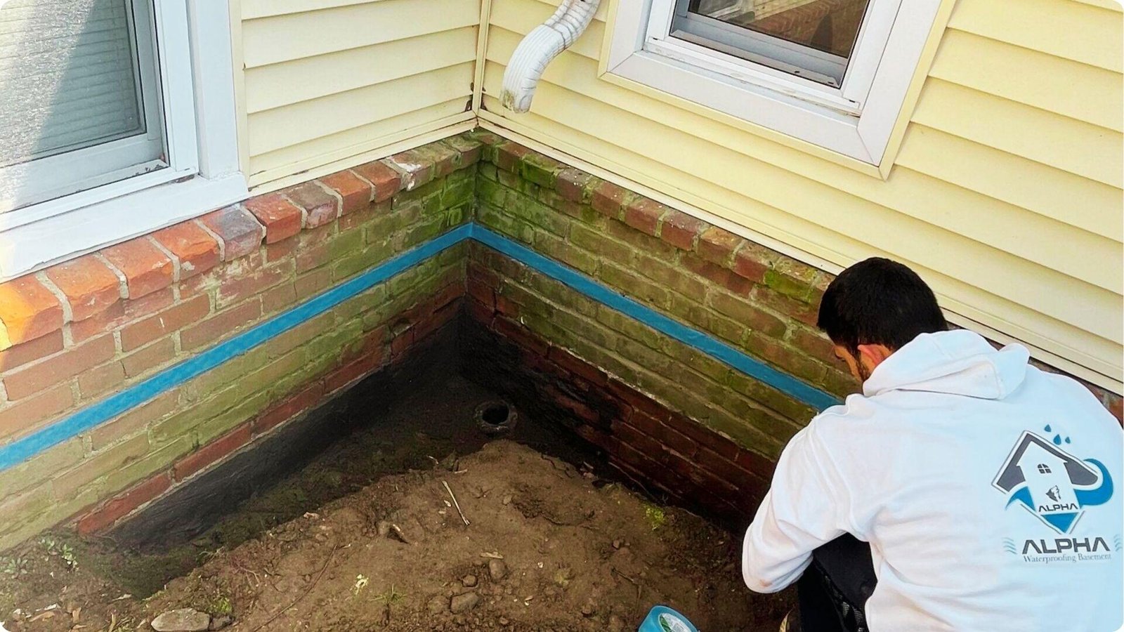 Worker from Alpha Waterproofing Basement installs blue sealant tape on brick foundation below siding.