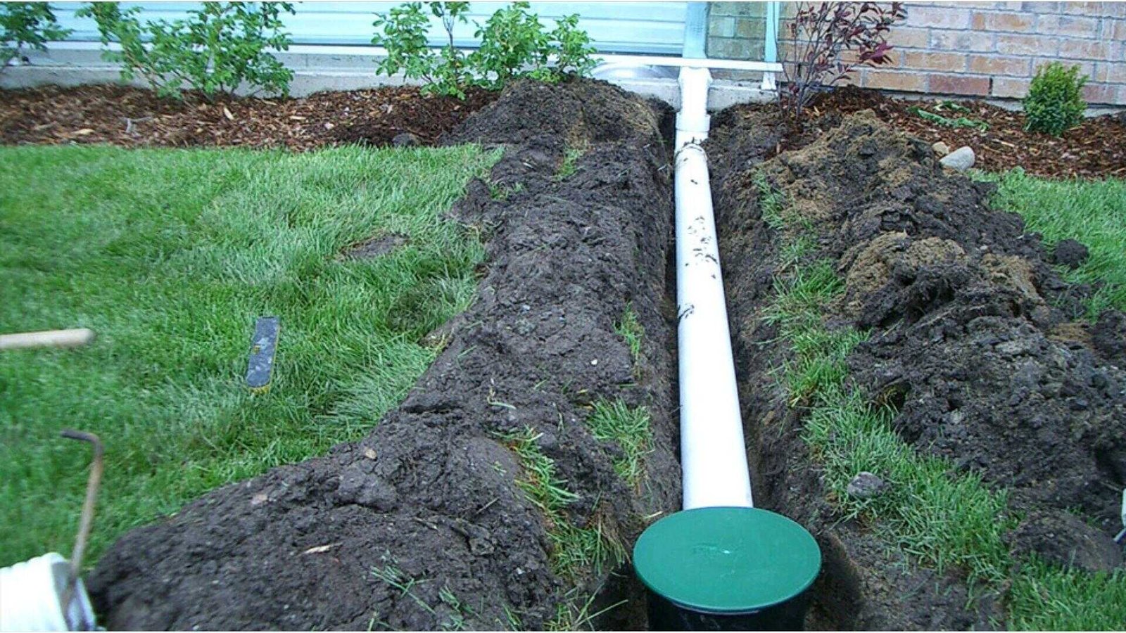 Exterior French drain installation with white PVC pipe in trench alongside house foundation to redirect groundwater