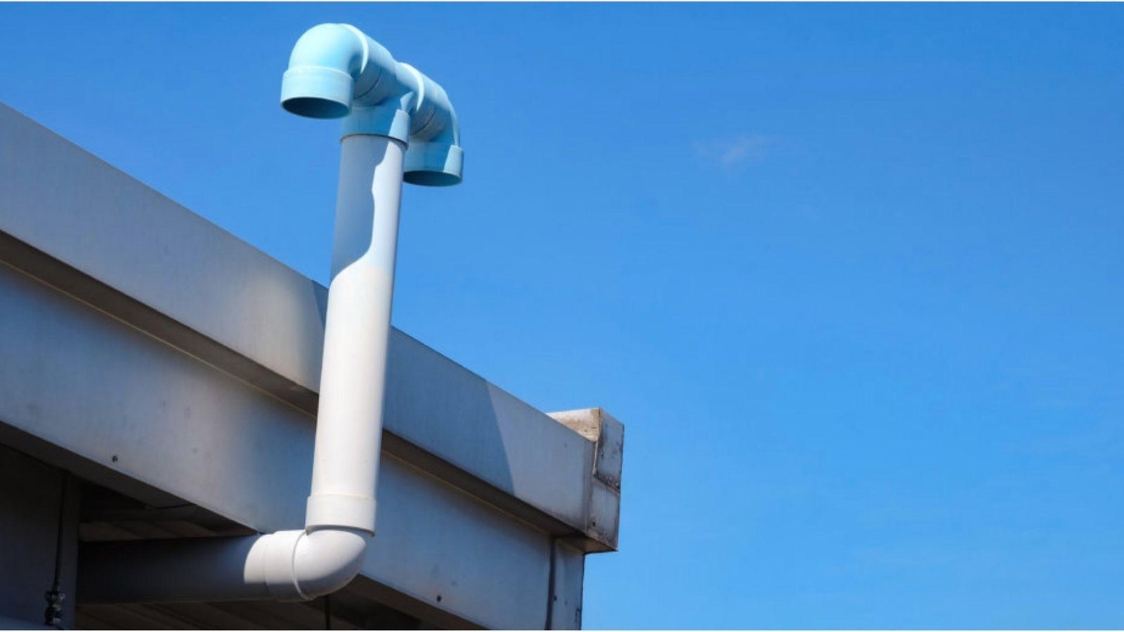 White PVC plumbing vent stack with blue pipe fittings extending above roofline against clear blue sky for basement bathroom ventilation