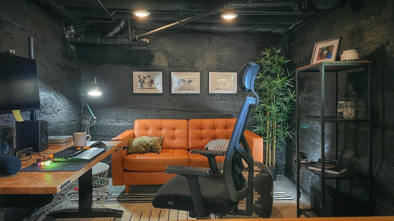 Industrial basement home office with orange sofa, desk setup, exposed pipes and concrete walls.