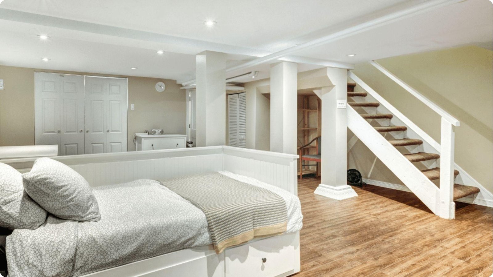 A bright English basement bedroom with a white daybed, wood flooring, and stairs leading up to the main floor.