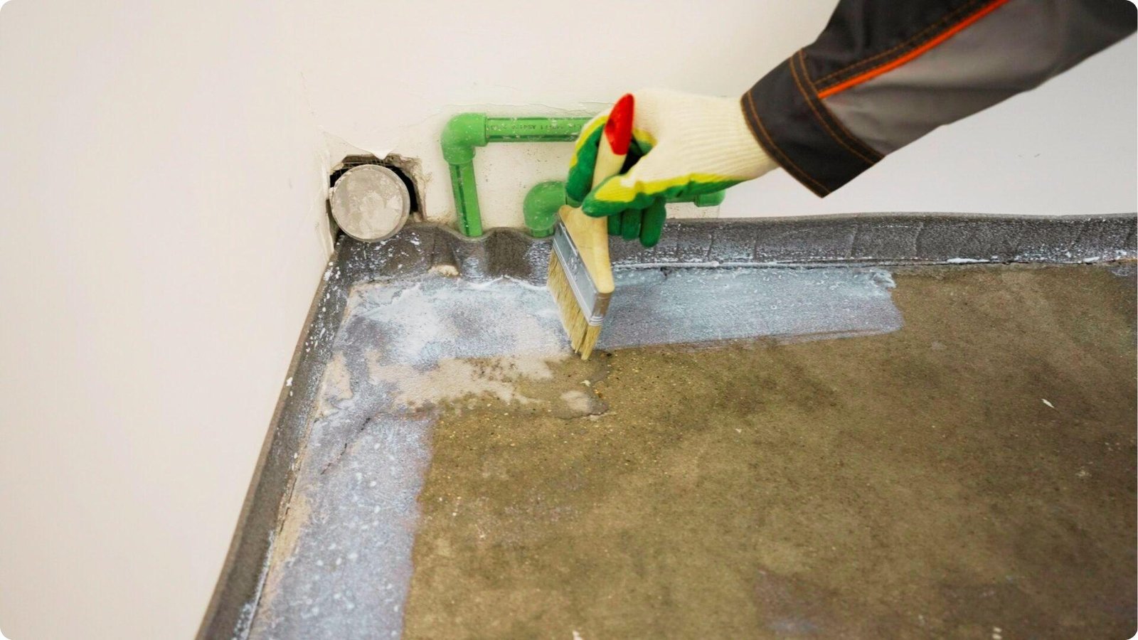 Gloved hand using a brush to apply liquid waterproofing membrane along the basement wall-floor joint near pipes.