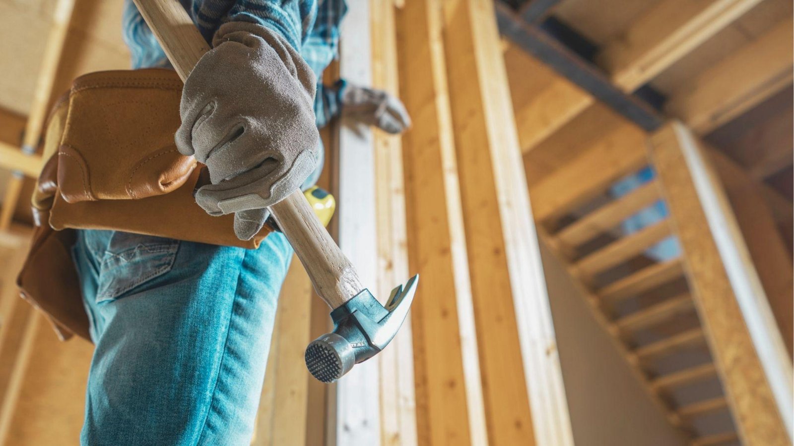 Low-angle view of a carpenter in gloves and a tool belt holding a hammer while framing interior basement walls.