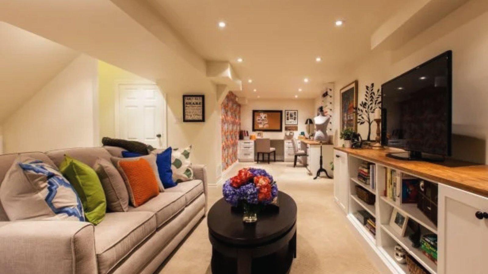 Basement craft room with beige sofa, colorful pillows, TV console, desks and mannequin for hobbies.
