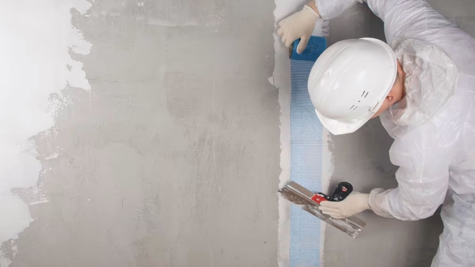 Worker applying fiberglass mesh tape over basement wall crack with trowel for structural repair.