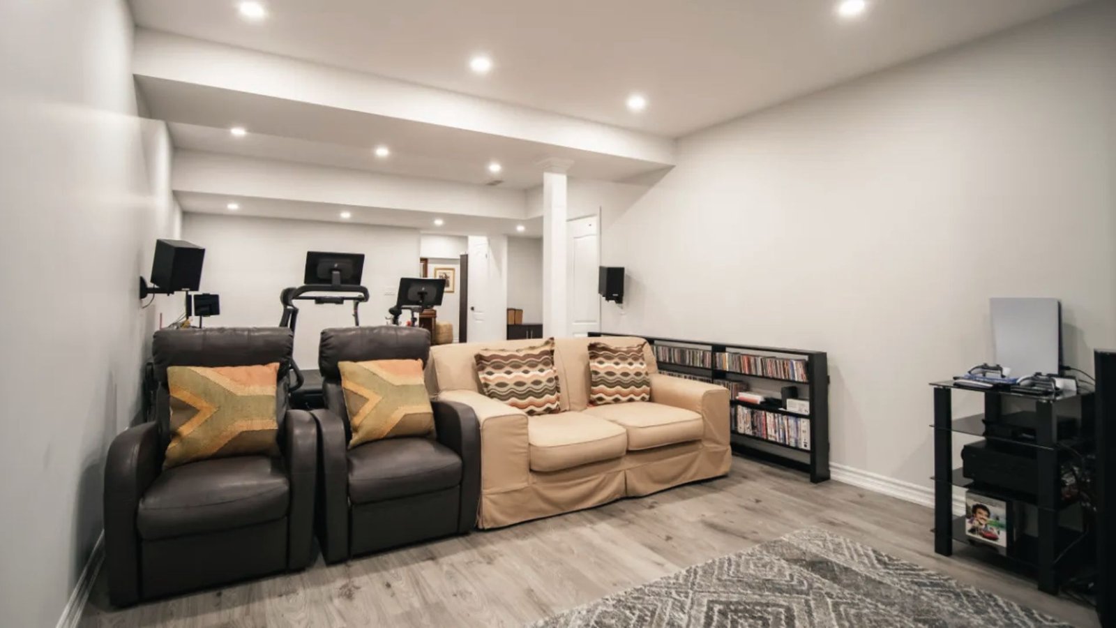Multipurpose basement with media seating, treadmill, game console, speakers and wood flooring.