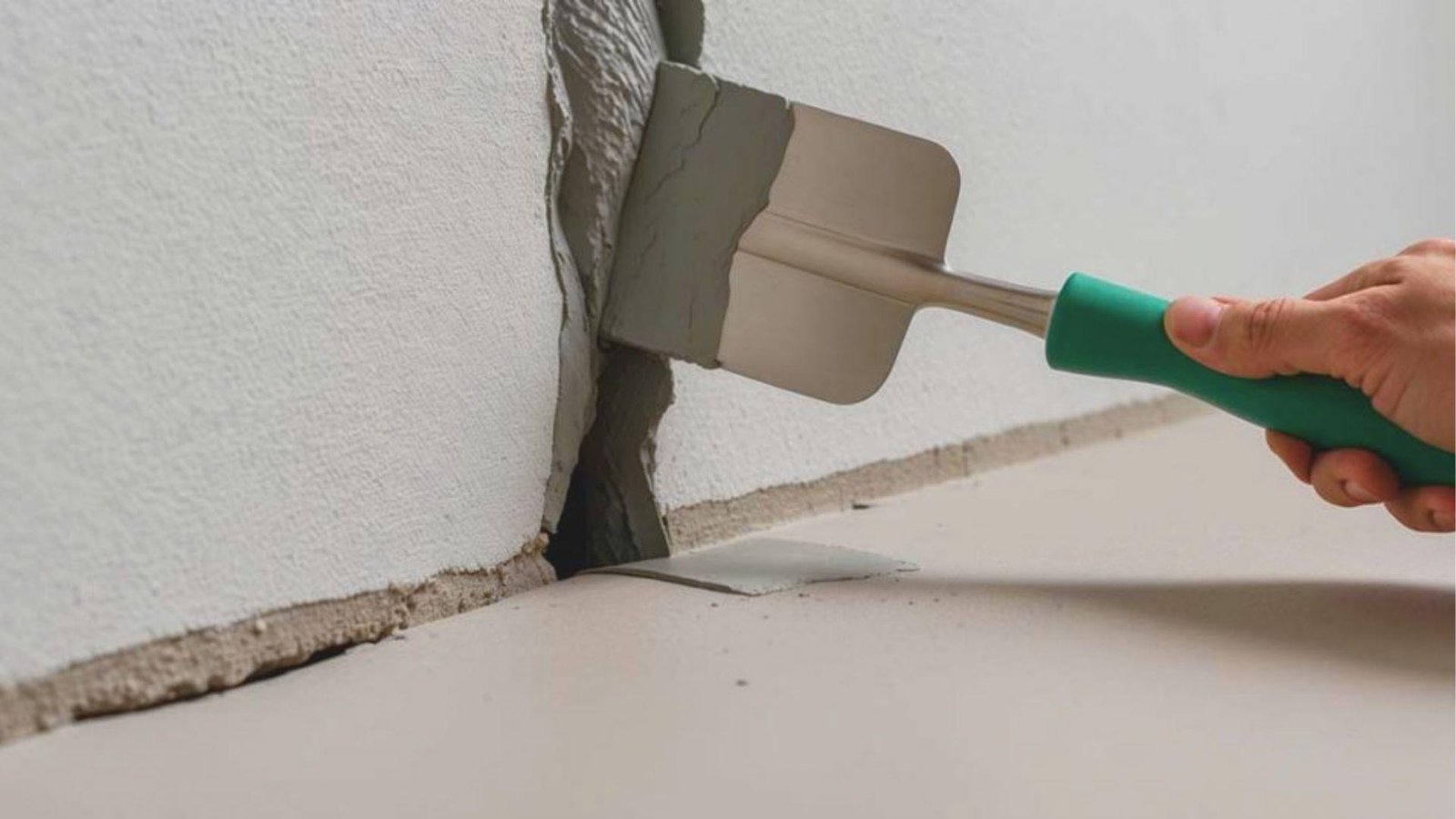 Close-up of a hand using a green-handled trowel to apply gray patching compound into a deep wall crack.