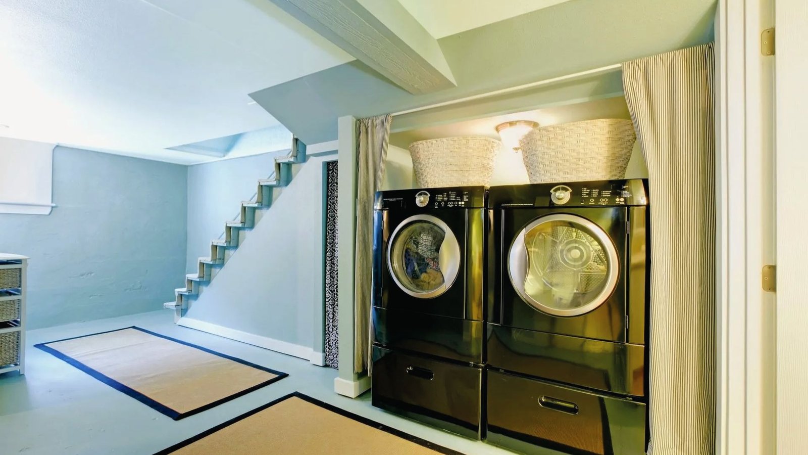 Basement laundry zone under stairs with black washer dryer, curtains, blue walls and rugs for utility.