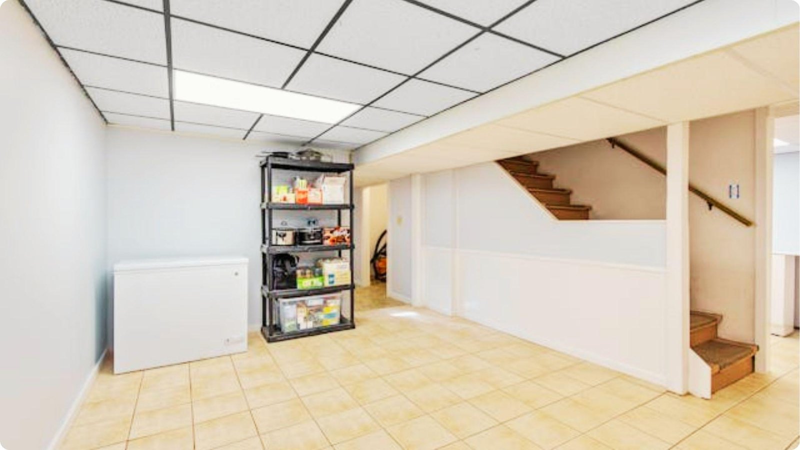Unfinished basement with drop ceiling, tile flooring, and storage shelving next to a staircase.