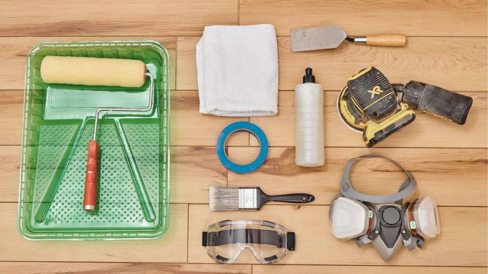 Flat lay of tools and safety gear for basement wall repair on wood flooring, including respirator, goggles, trowel, and sander.