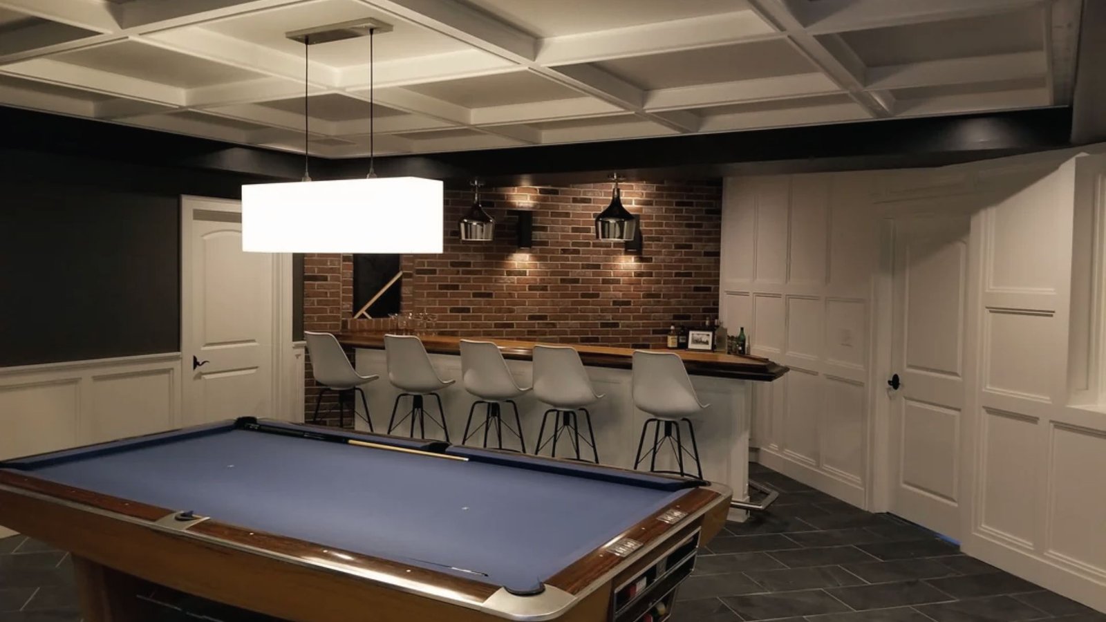 Industrial basement game room with pool table, brick bar, white stools and coffered ceiling.