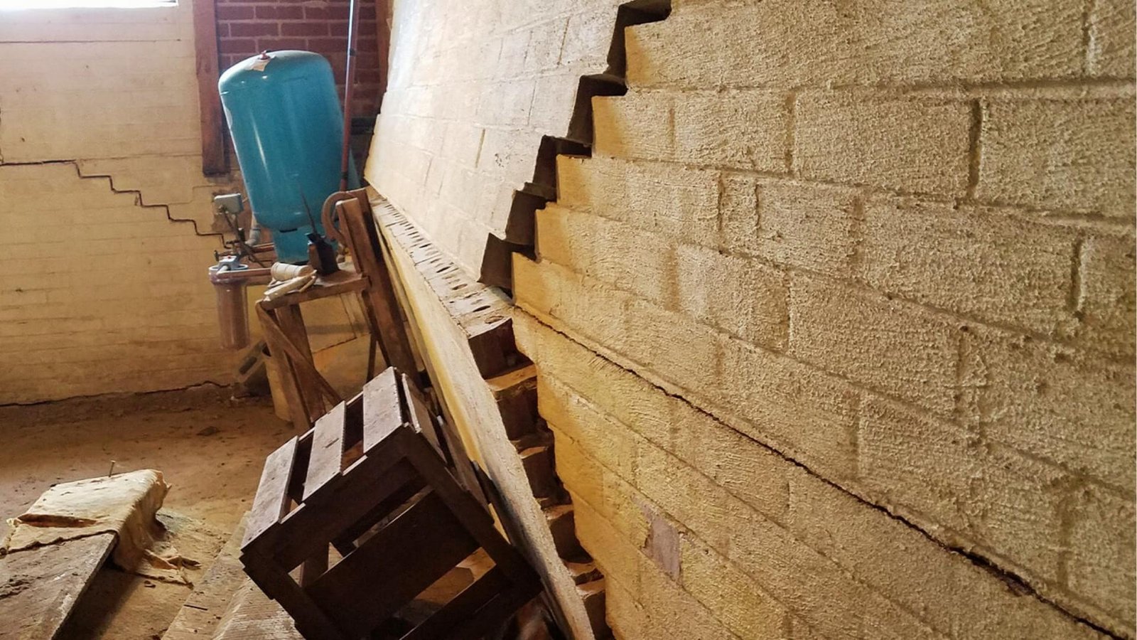 Severely bowed basement block wall with stair-step cracks and horizontal failure beside blue pressure tank, wooden crates, and leaning support beam.