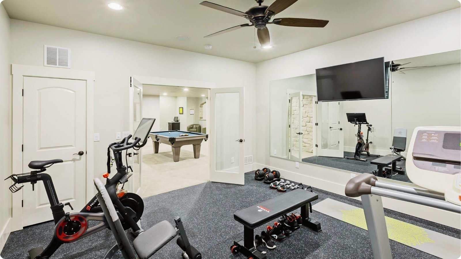 Basement home gym with a Peloton bike, treadmill, weights, and mirrored wall for workouts.