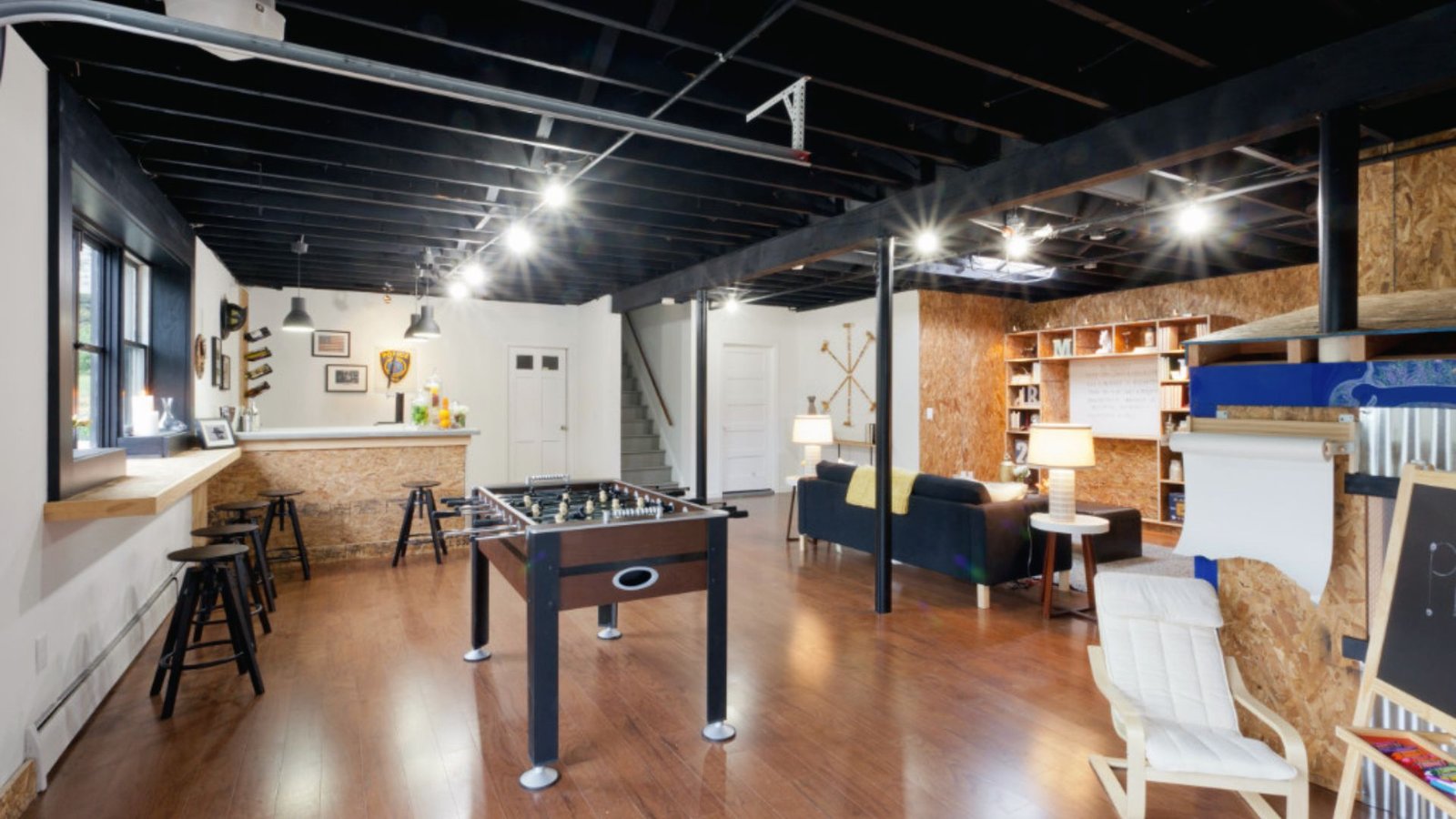 Industrial basement rec room with foosball table, wet bar, black ceiling, bookshelves and lounge sofa.